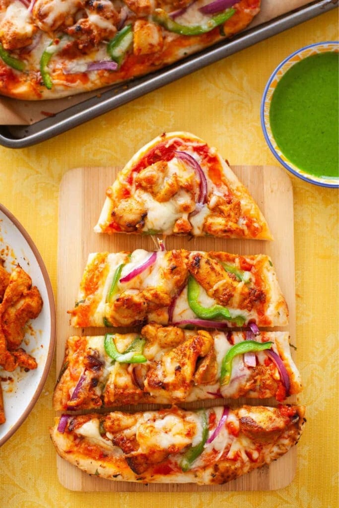 Naan pizza slices topped with tandoori-spiced paneer, melted mozzarella cheese, red onion, and fresh green peppers, served with cilantro chutney on a yellow background.