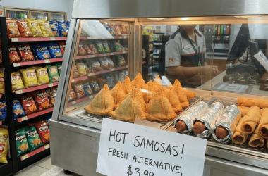 Hot case display featuring golden crispy samosas arranged in the front row at eye level, with a hand-written sign reading 'Hot Samosas! Fresh Alternative, $3.99' positioned in front of the warming cabinet.
